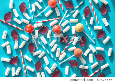 Composition of striped drinking straws, bright lollipops, airy marshmallows and pieces of candied fruit jelly lying in a certain order against blue background, directly above view Composition of striped drinking straws, bright lollipops, airy marshmallows and pieces of candied fruit jelly lying in a certain order against blue background, directly above view 89875336