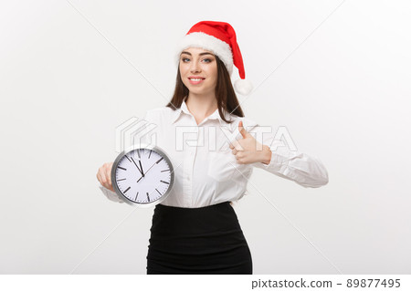 Time management concept - Young business woman with santa hat holding a clock and showing thump up isolated over white background. Time management concept - Young business woman with santa hat holding a clock and showing thump up isolated over white background. 89877495