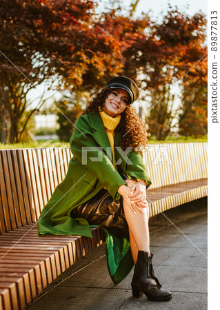 on a wooden bench in the park near the crimson trees sits a girl throwing her foot on her foot and folded on her knee her hands in beautiful stylish clothes 89877813