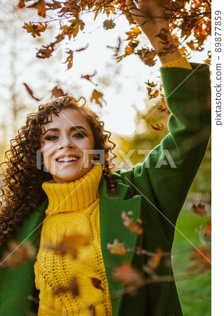 A positive girl with a beautiful white-toothed smile in warm autumn clothes stands under a tree with yellow leaves and touches them with her hand A positive girl with a beautiful white-toothed smile in warm autumn clothes stands under a tree with yellow leaves and touches them with her hand 89877859