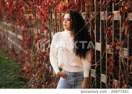 In the park at the wooden wall of twisted red ivy poses a girl in a white sweater and jeans 89877881