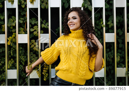 Against the background of the wooden wall behind which you can see the plants standing holding on to her hand a beautiful stylish girl in a bright yellow sweater and charmingly smiling 89877915