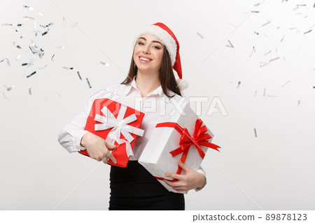 Business Concept - Beautiful young caucasian business woman with santa hat holding gift box with confetti celebration background. 89878123