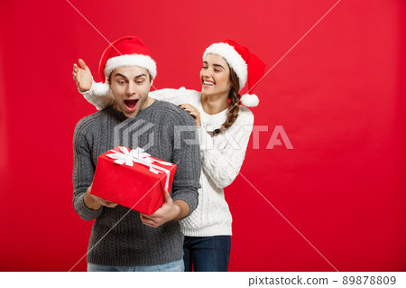 Christmas Concept - Young woman covering man's eyes with hand and giving surprise big gift. Isolated on Red background Christmas Concept - Young woman covering man's eyes with hand and giving surprise big gift. Isolated on Red background 89878809