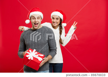Christmas Concept - Young woman covering man's eyes with hand and giving surprise big gift. Isolated on Red background Christmas Concept - Young woman covering man's eyes with hand and giving surprise big gift. Isolated on Red background 89878810