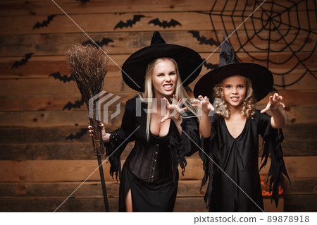 Halloween Concept - cheerful mother and her daughter in witch costumes celebrating Halloween posing with curved pumpkins over bats and spider web on Wooden studio background. Halloween Concept - cheerful mother and her daughter in witch costumes celebrating Halloween posing with curved pumpkins over bats and spider web on Wooden studio background. 89878918