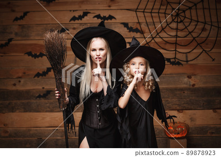 Halloween Concept - cheerful mother and her daughter in witch costumes celebrating Halloween doing silence gesture posing with curved pumpkins over bats and spider web on Wooden studio background. Halloween Concept - cheerful mother and her daughter in witch costumes celebrating Halloween doing silence gesture posing with curved pumpkins over bats and spider web on Wooden studio background. 89878923