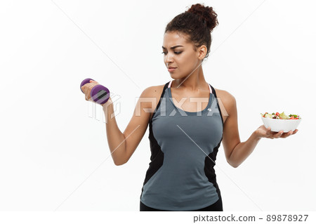 Healthy and Fitness concept - Beautiful sporty African American on diet holding dumbbell and fresh salad on hands. Isolated on white studio background. 89878927