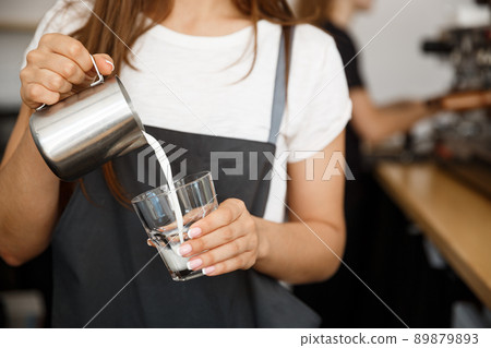 Coffee Business Concept - close-up lady barista in apron preparing and pouring milk in glass cup while standing at cafe. Coffee Business Concept - close-up lady barista in apron preparing and pouring milk in glass cup while standing at cafe. 89879893