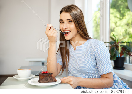 Young attractive Caucasian lady enjoy eating chocolate cake with hot coffee in modern coffee shop at noon. 89879921