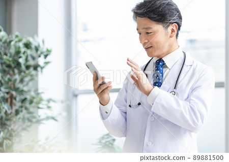 A doctor in a white coat standing by the window with a smartphone 89881500