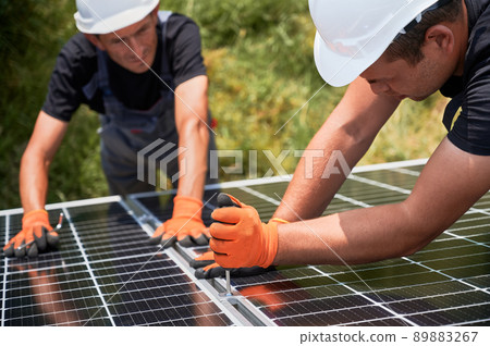 Male workers building photovoltaic solar panel system outdoors. Men engineers placing solar module on metal rails, wearing construction helmets and work gloves. Renewable and ecological energy. Male workers building photovoltaic solar panel system outdoors. Men engineers placing solar module on metal rails, wearing construction helmets and work gloves. Renewable and ecological energy. 89883267