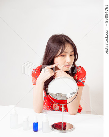 Portrait of a young asian woman in traditional cheongsam qipao dress looks at mirror and does skin care isolated on white background. Portrait of a young asian woman in traditional cheongsam qipao dress looks at mirror and does skin care isolated on white background. 89884763