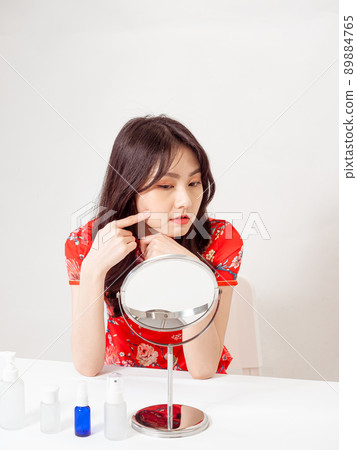 Portrait of a young asian woman in traditional cheongsam qipao dress looks at mirror and does skin care isolated on white background. Portrait of a young asian woman in traditional cheongsam qipao dress looks at mirror and does skin care isolated on white background. 89884765