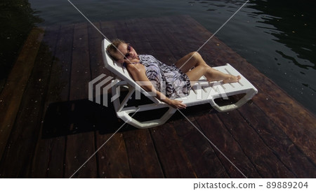 Woman lie on a sunbed in sunglasses and a boho silk shawl. Girl rest on a flood wood underwater pier. The pavement is covered with water in the lake. Woman lie on a sunbed in sunglasses and a boho silk shawl. Girl rest on a flood wood underwater pier. The pavement is covered with water in the lake. 89889204