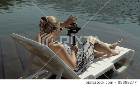 Woman shooting on handheld film gimbal stabilization for smartphone. Girl lie sunbed on pier make selfie. Blogger broadcast video vlogging. Woman shooting on handheld film gimbal stabilization for smartphone. Girl lie sunbed on pier make selfie. Blogger broadcast video vlogging. 89889277