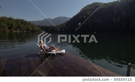 Woman lie on a sunbed in sunglasses and a boho silk shawl. Girl rest on a flood wood underwater pier. The pavement is covered with water in the lake. 89889318