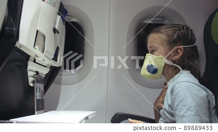 Little girl caucasian at plane with wearing protective mask. Child baby tourist at aircraft with respirator play at smartphone mobile. Covid-19. 89889433
