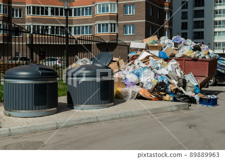 Crowded garbage cans in the courtyard of a residential apartment building. Garbage sorting containers. Trash is scattered on the sidewalk. Crowded garbage cans in the courtyard of a residential apartment building. Garbage sorting containers. Trash is scattered on the sidewalk. 89889963
