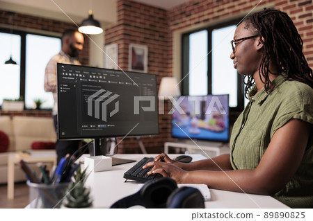Woman programmer working remote writing code using computer while boyfriend is relaxing watching tv. African american freelancer developing software on pc sitting at desk in home living room 89890825