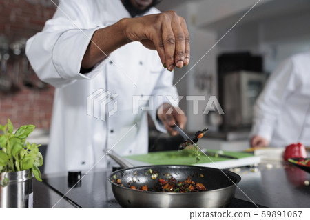 Master chef seasoning dish prepared for food contest held at fine dining restaurant. Head cook throwing fresh chopped herbs in pan to improve taste of meal while in professional kitchen. Master chef seasoning dish prepared for food contest held at fine dining restaurant. Head cook throwing fresh chopped herbs in pan to improve taste of meal while in professional kitchen. 89891067