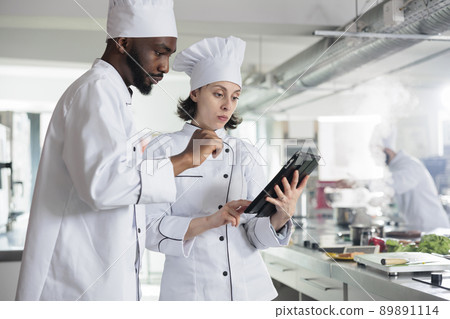 Professional chefs with wireless touchscreen device preparing ingredients for food contest held at restaurant. Gourmet cuisine experts with modern tablet searching for dinner dish ideas on internet. Professional chefs with wireless touchscreen device preparing ingredients for food contest held at restaurant. Gourmet cuisine experts with modern tablet searching for dinner dish ideas on internet. 89891114