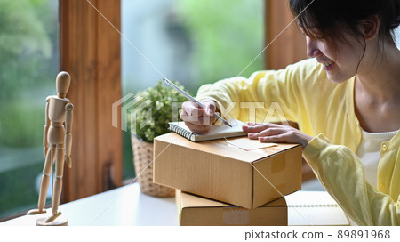 Focused young woman online seller writing address on cardboard box while preparing parcel boxes of product for delivery 89891968