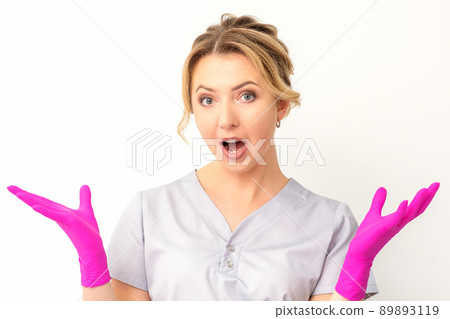 Body language. Young surprised Caucasian woman doctor wearing gloves gesturing with her hands, spread his arms having shocked expression opening her mouth against the white background 89893119