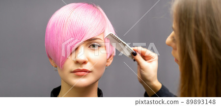 The female hairdresser is combing the dyed pink short hair of the young woman against the dark wall 89893148