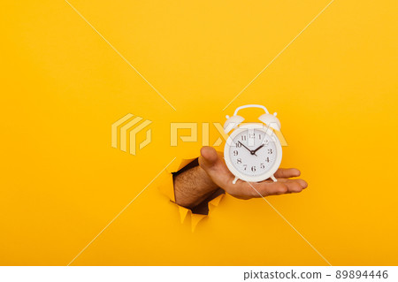 Hand holds alarm clock through a paper hole in yellow background with copy space 89894446