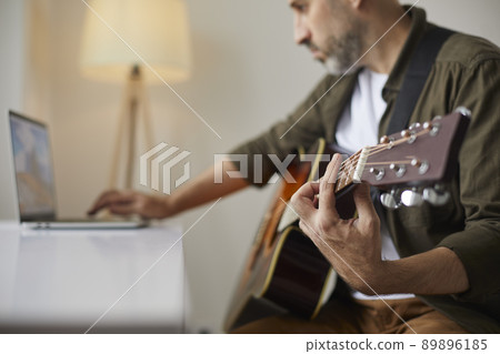 Man with acoustic guitar clamps desired chord while watching online lesson on laptop. 89896185
