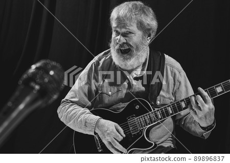Black and white portrait of stylish old man, rock musician playing guitar at music studio interior. Rehearsal base Concept of art, music, style, older generation, vintage 89896837