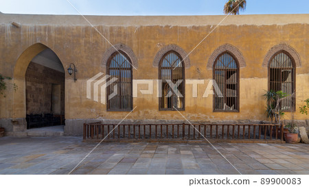 Wooden arched window with wrought iron grid over yellow stone bricks wall and arched passage 89900083