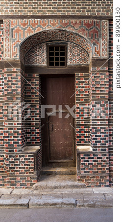 Grunge narrow wooden decorated arched gate with inner small door on wall with black and red bricks 89900130