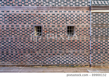 Wall with black and red bricks with white seam and two small windows with metal bars 89900132