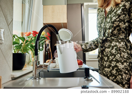 Woman pouring clean filtered water from faucet into electric kettle for boiling water at kitchen 89902188