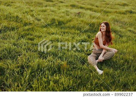 The sunset light of a summer evening illuminates a beautiful young redheaded woman sitting on the grass in green pants and an orange top and smiling at the sun The sunset light of a summer evening illuminates a beautiful young redheaded woman sitting on the grass in green pants and an orange top and smiling at the sun 89902237