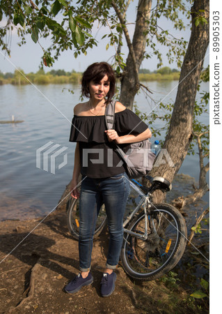 An adult woman with a bicycle stopped on the shore of a lake enjoying nature An adult woman with a bicycle stopped on the shore of a lake enjoying nature 89905303