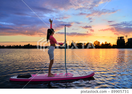 Fitness young woman standing on sup board with long paddle in hands and enjoying amazing summer sunset over lake. Outdoors activity during leisure time. 89905731
