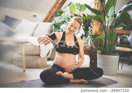 Young happy and cheerful beautiful pregnant woman taking selfie with her mobile phone while staying fit, sporty and active on her maternity leave. Motherhood, pregnancy, yoga concept. 89907109