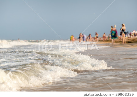 Scenic view sand shore of sea or ocean waves on sunny beach of summer resort crowded with many people relaxing on hot day. Tourism vacation concept Scenic view sand shore of sea or ocean waves on sunny beach of summer resort crowded with many people relaxing on hot day. Tourism vacation concept 89908359