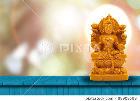 The statue of Mahalakshmi carved in brown wood on a wooden table has a natural bokeh background with a powerful light behind the head The statue of Mahalakshmi carved in brown wood on a wooden table has a natural bokeh background with a powerful light behind the head 89909599
