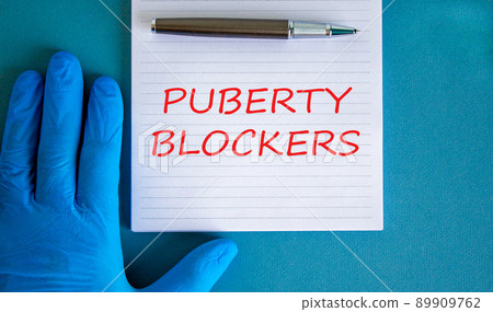 Medical and puberty blockers symbol. Words 'puberty blockers' on white card. Doctor hand in blue glove and metallic pen. Beautiful white background. Copy space. Medical and puberty blockers concept. Medical and puberty blockers symbol. Words 'puberty blockers' on white card. Doctor hand in blue glove and metallic pen. Beautiful white background. Copy space. Medical and puberty blockers concept. 89909762