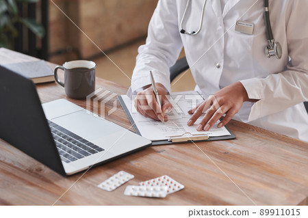Unrecognizable female doctor wearing white coat sitting at desk in loft office writing something in papers Unrecognizable female doctor wearing white coat sitting at desk in loft office writing something in papers 89911015