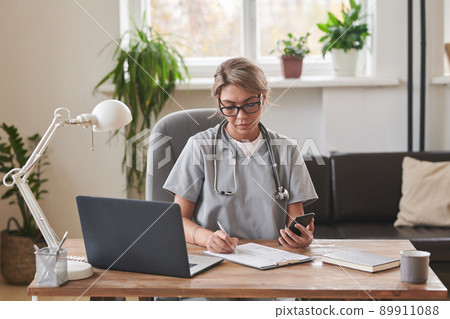 Horizontal medium shot of young adult female medical specialist sitting at desk in loft office watching something on smartphone and making notes 89911088