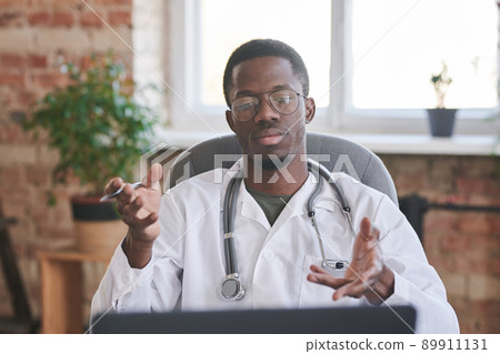 Medium portrait of modern young adult doctor wearing white coat and eyeglasses sitting in loft office room talking to patient online 89911131