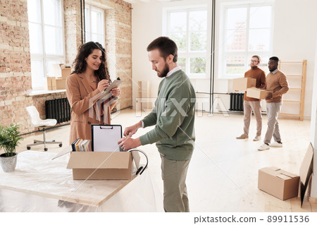 Smiling young woman checking list while her colleague unpacking moving box of documents in new office Smiling young woman checking list while her colleague unpacking moving box of documents in new office 89911536