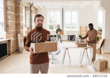 Portrait of content handsome young manager in turtleneck holding closed stuff box against colleagues setting up new office Portrait of content handsome young manager in turtleneck holding closed stuff box against colleagues setting up new office 89911538
