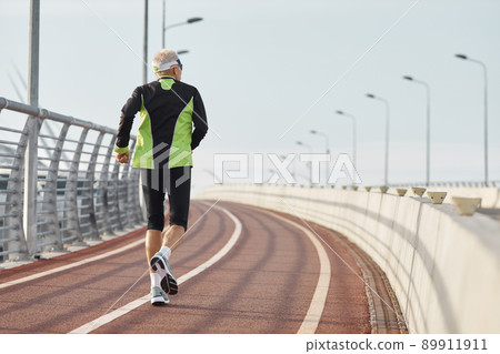 Horizontal back view shot of senior man wearing black and green tracksuit running along track 89911911