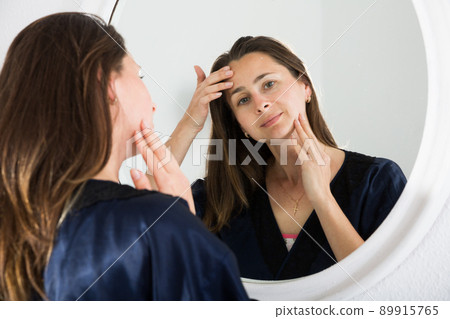 Portrait of adult woman looking at mirror and touching face in bedroom Portrait of adult woman looking at mirror and touching face in bedroom 89915765
